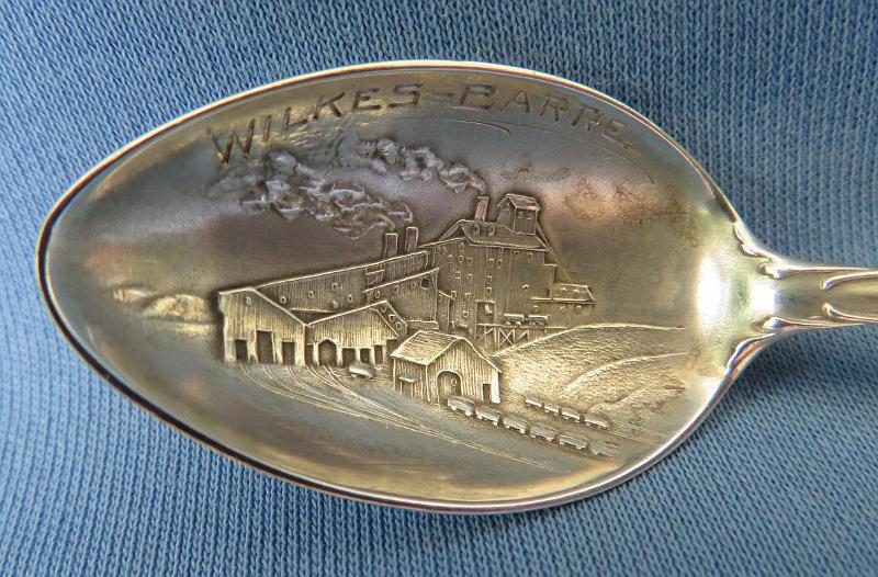 Souvenir Mining Spoon Wilkes-Barre Bowl.JPG - SOUVENIR MINING SPOON WILKES-BARRE PA - Sterling silver souvenir demitassespoon, 4 in. long, bowl engraved with colliery buildings and marked WILKES-BARRE, handle marked with image of William Penn and Liberty Bell with Pennsylvania, reverse marked Sterling and maker’s mark of Mechanics Sterling Co. (Attleboro, MA 1896 - ?), reverse handle marked with pick and shovel along with state seal  [Wilkes-Barre is in the center of the Wyoming Valley anthracite coal region in northeastern Pennsylvania.  Founded in 1769, it was originally named Wyoming but renamed later in honor of two British members of parliament, John Wilkes and Col. Issac Barre, who defended the American colonies in parliamentary debates.  In 1818, Wilkes-Barre was incorporated as a borough, with a city charter following in 1871.  Coal mining was the most important element of the city’s economy.   Hundreds of thousands of immigrants flocked to the city; they were seeking jobs in the numerous mines and collieries that sprang up.  Wilkes-Barre’s population exploded as the city became a center of supply to support these mines.  In 1914 employment at the anthracite mines reached a maximum of 180,000 workers.  Anthracite production peaked in 1917 at over 100 million tons with 776 mines in operation.  The anthracite industry went into steady decline after World War I.  The primary reason was competition from abundant supplies of lower cost oil and gas.  A large drop in anthracite production occurred during the Depression with only a small bounce-back during World War II.  The earlier downward trend continued after the War.  In 1959, The Knox Mine near Wilkes-Barre broke through the bottom of the Susquehanna River, flooding the underground mines and ending deep coal  production in the area.]